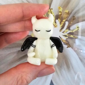 May include: A small, off-white figurine of a seated devil with closed eyes, small horns, and black wings. The figurine has black stripes on its arms and is held between two fingers.