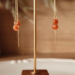 May include: Gold-toned dangle earrings featuring two orange-red, orb-shaped beads. The earrings hang from a delicate chain and a thin, gold bar. The earrings are displayed on a gold stand with a wooden base.
