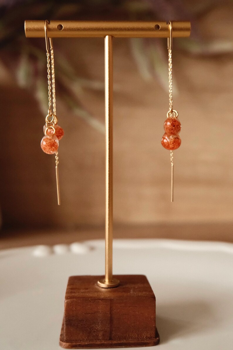 May include: Gold-toned dangle earrings featuring two orange-red, orb-shaped beads. The earrings hang from a delicate chain and a thin, gold bar. The earrings are displayed on a gold stand with a wooden base.