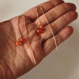 May include: A pair of gold-colored threader earrings. Each earring features a small cluster of three red-orange, speckled beads. The earrings are displayed against a neutral background.