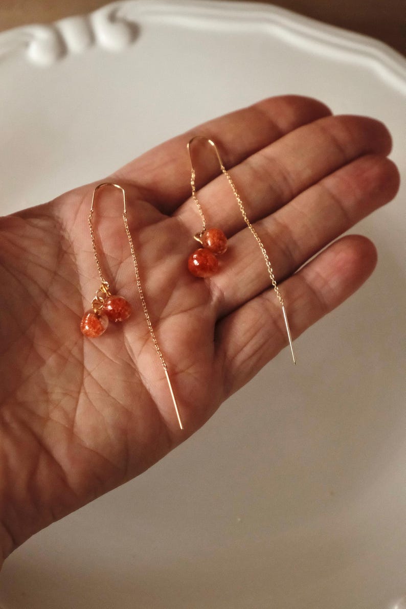 May include: A pair of gold-colored threader earrings. Each earring features a small cluster of three red-orange, speckled beads. The earrings are displayed against a neutral background.