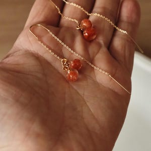 May include: Delicate gold-tone threader earrings with small, round, orange-red beads. The earrings are displayed against a warm, neutral background, showcasing their simple yet elegant design. These earrings are a stylish accessory.