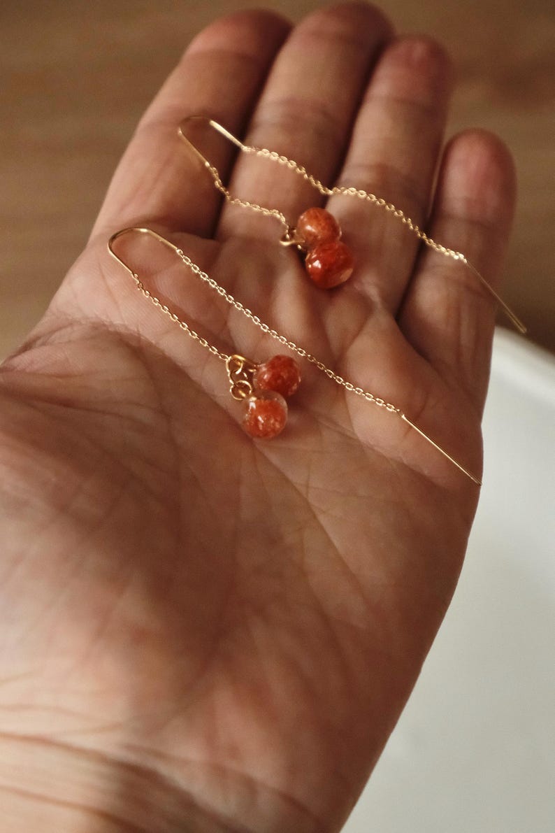 May include: Delicate gold-tone threader earrings with small, round, orange-red beads. The earrings are displayed against a warm, neutral background, showcasing their simple yet elegant design. These earrings are a stylish accessory.