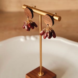 Könnte beinhalten: Ohrringe auf einem goldenen Ständer. Die Ohrringe haben kleine, dunkelrote, tropfenförmige Perlen, die an goldenen Ketten hängen. Die Ohrringe sind an Holzscheiben befestigt. Der Ständer hat einen Holzsockel.