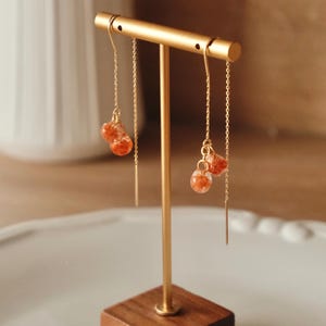 May include: Gold-colored dangle earrings with small, round, orange-red beads. The earrings hang from a gold chain and are displayed on a gold stand with a wooden base. The background is blurred.