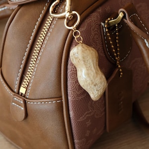Könnte beinhalten: Braune Lederhandtasche mit goldenem Reißverschluss und Beschlägen. Ein Erdnuss-förmiger Anhänger hängt am Reißverschluss. Die Tasche hat weiße Nähte und ein gemustertes Stoffpaneel. Die Abmessungen der Tasche betragen ca. 30 cm lang, 20 cm hoch und 15 cm breit.