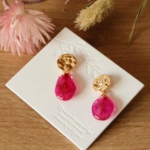 Könnte beinhalten: Goldfarbene Ohrringe mit strukturiertem, kreisförmigem Oberteil und einem tropfenförmigen, magenta-farbenen Anhänger. Die Ohrringe werden auf einer weißen quadratischen Karte mit floralen Akzenten und Text präsentiert. Der Hintergrund umfasst getrocknete Blumen und eine Holzoberfläche.