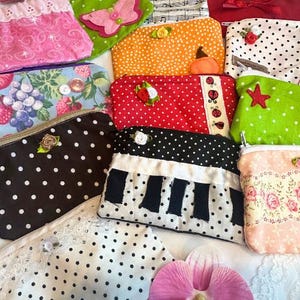Puede incluir: Una colecci&oacute;n de bolsas de tela hechas a mano en varios colores y patrones. Los dise&ntilde;os incluyen lunares, estampados florales y detalles aplicados como mariposas, estrellas y notas musicales. Cada bolsa tiene cierre de cremallera y adornos.