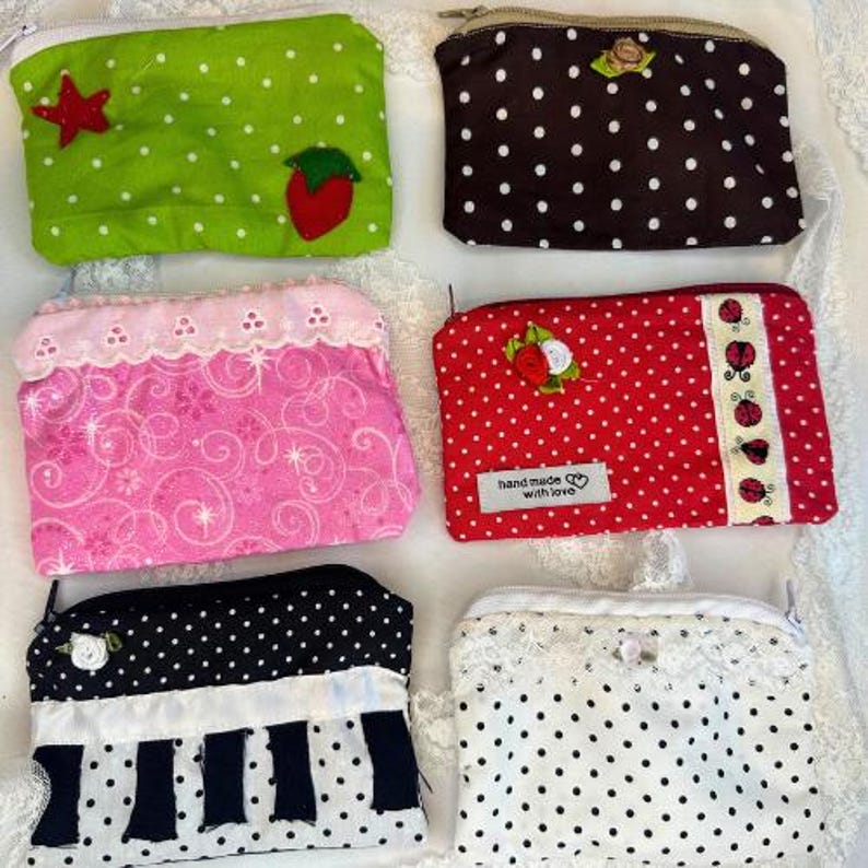 Puede incluir: Una colecci&oacute;n de seis monederos con cremallera de tela hechos a mano. Los monederos presentan varios patrones, incluyendo lunares, dise&ntilde;os florales y adornos aplicados como una fresa y una estrella. La paleta de colores incluye verde, marr&oacute;n, rosa, rojo, negro y blanco.