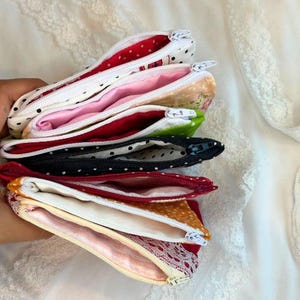 Puede incluir: Una pila de estuches con cremallera abiertos en varios colores y patrones. Los estuches incluyen blanco con lunares negros, rosa, floral, verde, negro con lunares blancos y rojo con lunares blancos. Los interiores est&aacute;n forrados con telas a juego.