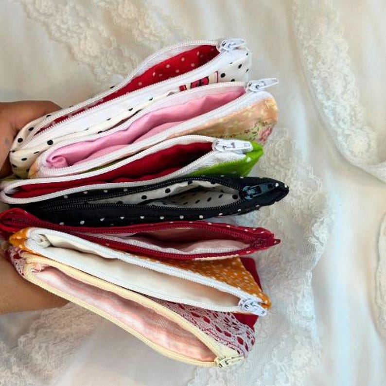 Puede incluir: Una pila de estuches con cremallera abiertos en varios colores y patrones. Los estuches incluyen blanco con lunares negros, rosa, floral, verde, negro con lunares blancos y rojo con lunares blancos. Los interiores est&aacute;n forrados con telas a juego.