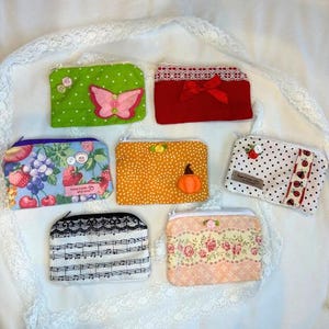Puede incluir: Una colecci&oacute;n de monederos con cremallera de tela hechos a mano en varios colores y patrones. Los dise&ntilde;os incluyen lunares, estampados florales, una mariposa, una calabaza y notas musicales. Cada monedero tiene un elemento decorativo &uacute;nico, como encaje o un lazo.