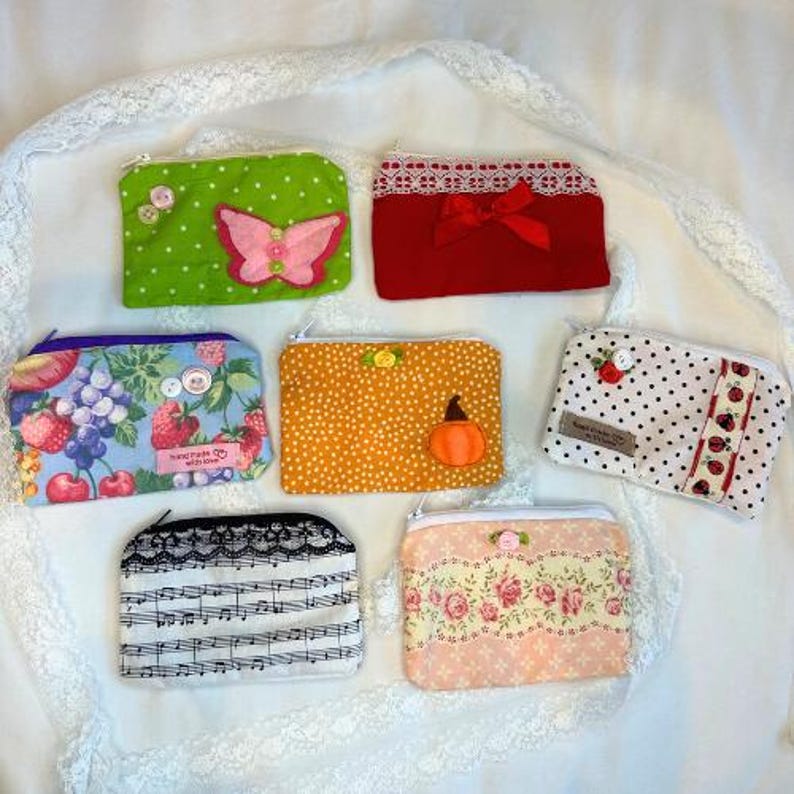 Puede incluir: Una colecci&oacute;n de monederos con cremallera de tela hechos a mano en varios colores y patrones. Los dise&ntilde;os incluyen lunares, estampados florales, una mariposa, una calabaza y notas musicales. Cada monedero tiene un elemento decorativo &uacute;nico, como encaje o un lazo.