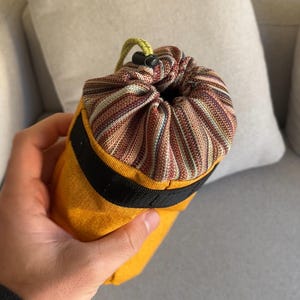 Puede incluir: Una bolsa cilíndrica amarilla con una franja negra y un cierre de cordón a rayas. La bolsa se sostiene en una mano y tiene un cordón verde y amarillo. La parte superior de la bolsa es un patrón a rayas marrón, rojo y blanco.