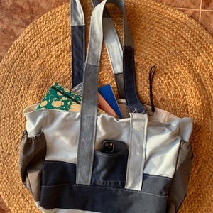 Puede incluir: Un bolso tote de mezclilla con múltiples tonos de azul y gris. El bolso tiene dos correas largas y un bolsillo frontal. El bolso está lleno de objetos, incluyendo un teléfono y un cuaderno. El bolso está sobre una estera redonda tejida.