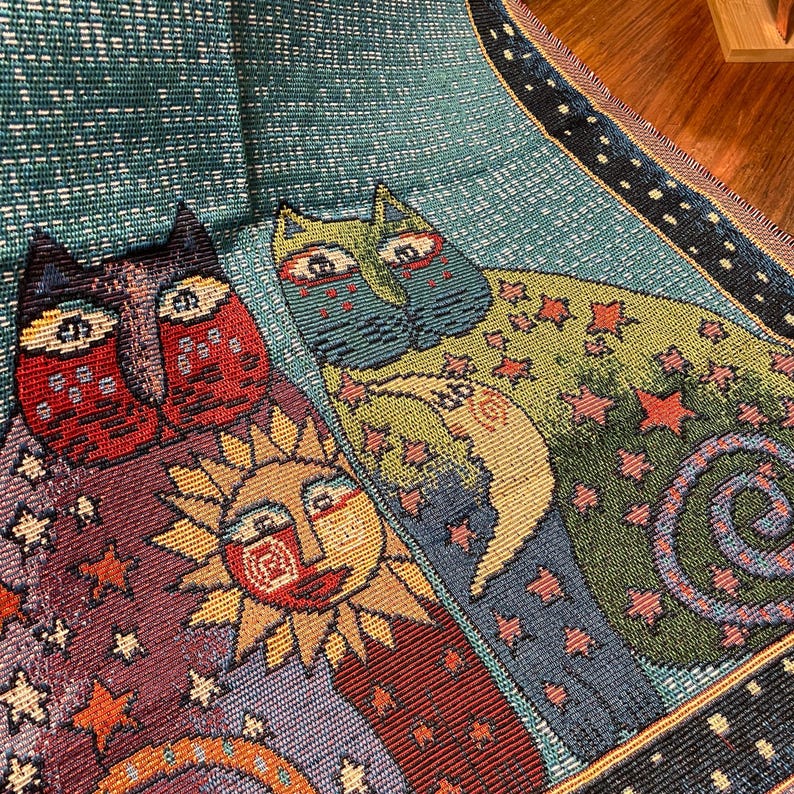 以下が含まれることがあります： 3匹の風変わりな猫、太陽、星が描かれたタペストリー。猫は青、緑、赤の色合いで、太陽は黄色とオレンジ色。背景はターコイズブルーで、白い星が散りばめられています。