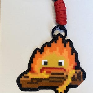 以下が含まれることがあります： オレンジと黄色の色合いの炎のキャラクター、黒い目、赤い口が茶色の丸太の上に乗っているピクセルアートスタイルのキーホルダー。デザインには黒いグリッターの縁取りがあり、黒いリングを介して赤いパラコードストラップに取り付けられています。