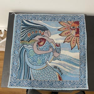 以下が含まれることがあります： 青い波模様のタペストリーに、2匹の魚を抱えた人魚が描かれています。人魚は長い青い髪で、青い波に囲まれています。タペストリーの右上には太陽が描かれています。タペストリーは青い縁取りがされており、ペイズリー柄が施されています。右下には「sea-maid」という文字が書かれています。