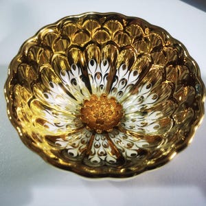 Könnte beinhalten: Eine goldene und weiße Keramikschale mit einem dekorativen Blumendesign. Die Schale hat eine erhabene, strukturierte Oberfläche und einen gewellten Rand.