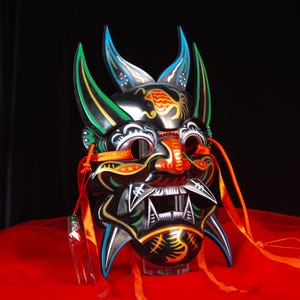 May include: A black decorative mask with a fierce expression, featuring sharp teeth, red accents, and colourful details. The mask has long, pointed horns and is adorned with orange ribbons. The mask is displayed on a red surface.