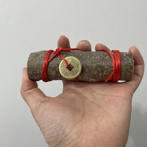 Puede incluir: Un trozo cilíndrico de madera con una moneda de bronce y una cuerda roja enrollada. La moneda tiene caracteres chinos. La madera se sostiene en una mano.