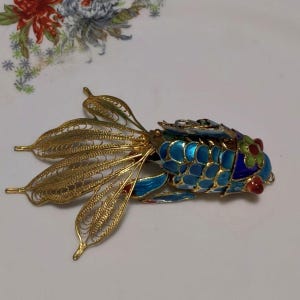 以下が含まれることがあります： 金色の金属製の魚のブローチ。複雑なフィリグリー細工の尾びれと、青、緑、赤のエナメル質の鱗で装飾された胴体が特徴です。頭部は鮮やかな青と緑のエナメル模様で、目の近くに小さな赤いアクセントが施されています。