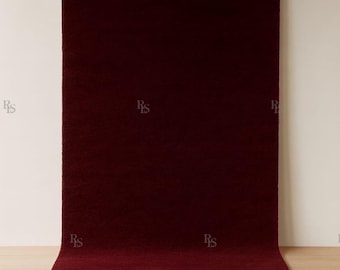 May include: A rectangular, burgundy-colored rug with a soft, textured surface. The rug is displayed on a light-colored wooden floor, with a neutral-toned wall in the background. The rug is a solid color and appears to be made of a plush material.