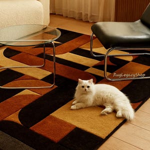 Pode incluir: Um tapete moderno com um padr&atilde;o geom&eacute;trico em tons de castanho, preto e dourado. Um gato branco est&aacute; deitado no tapete. O tapete apresenta um design contempor&acirc;neo com formas curvas e retangulares. O texto "RugLegacyShop" &eacute; vis&iacute;vel.