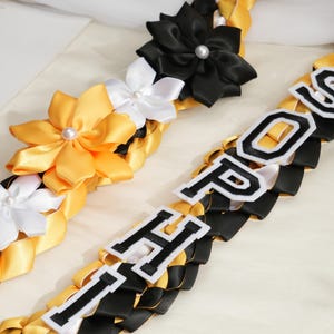 May include: Braided leis in black, gold, and white, adorned with satin flowers and the letters "HOP" and "POS" in a collegiate font. The leis are likely for a graduation or celebratory event.