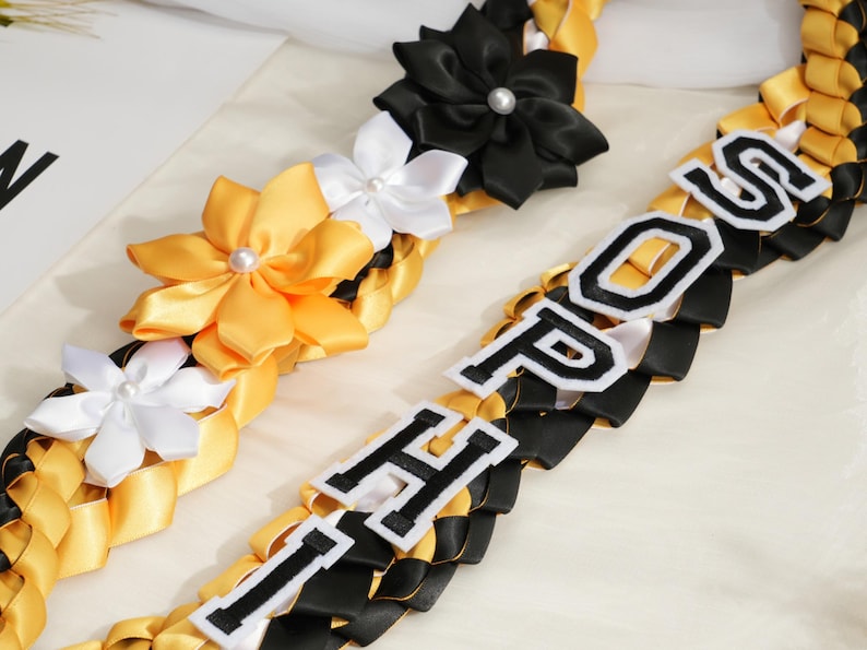 May include: Braided leis in black, gold, and white, adorned with satin flowers and the letters "HOP" and "POS" in a collegiate font. The leis are likely for a graduation or celebratory event.
