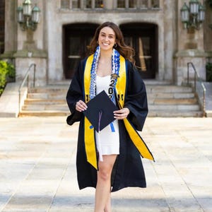 May include: A person in a graduation gown, holding a mortarboard and wearing a sash with the year 2023. The gown is black with yellow trim, and the sash is blue and white. The person is wearing a white dress and nude heels.