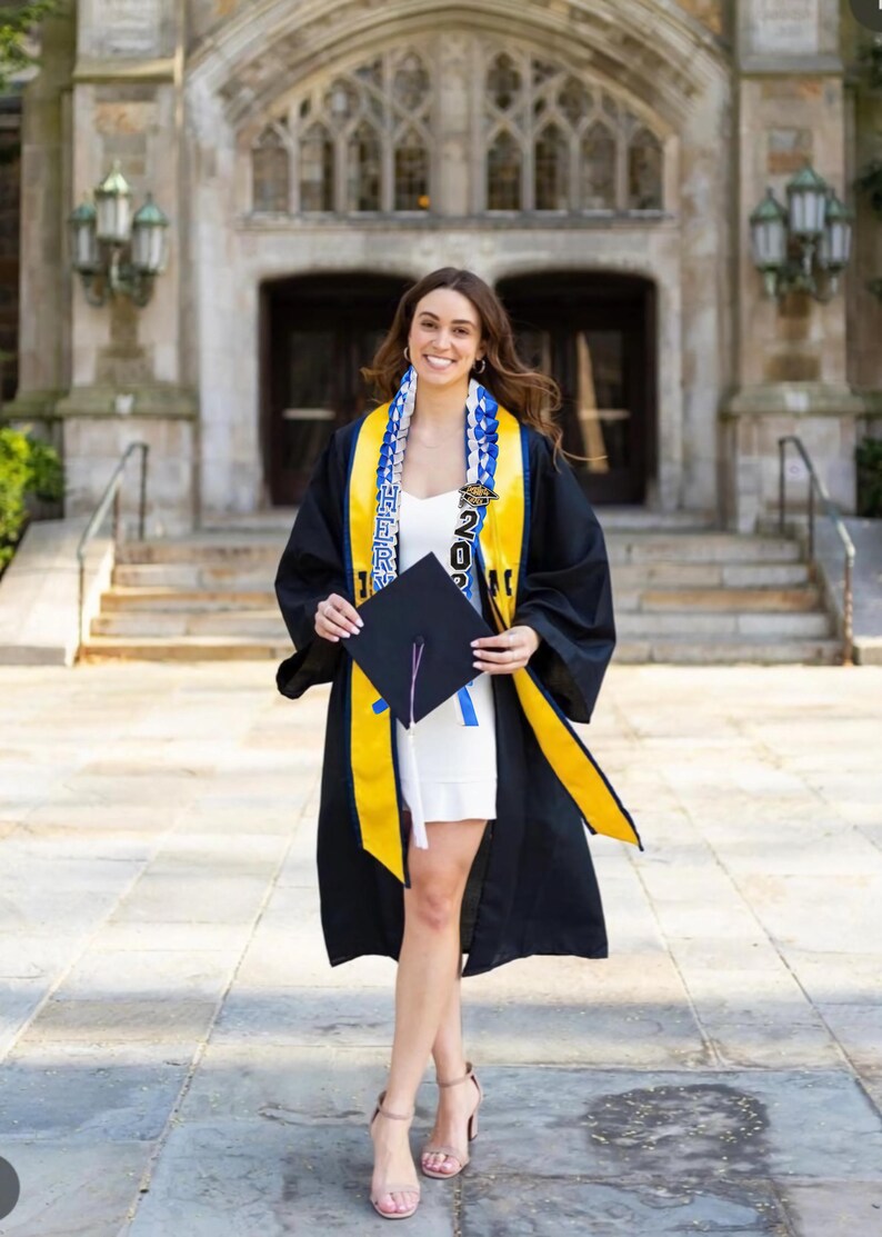 May include: A person in a graduation gown, holding a mortarboard and wearing a sash with the year 2023. The gown is black with yellow trim, and the sash is blue and white. The person is wearing a white dress and nude heels.