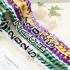 May include: Braided graduation leis in green, white, purple, and gold. Each lei features the year "2025" and a graduation cap. One lei has the name "Aaron". The leis are displayed on a light-colored surface.