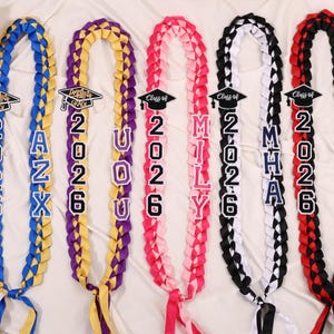 May include: Braided graduation leis in blue, yellow, purple, pink, black, and red. Each lei features the year "2026" and a name or initials, with a graduation cap icon. The leis are made of ribbon and are displayed on a white surface.