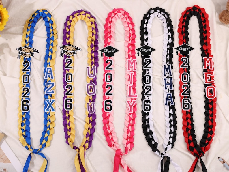 May include: Braided graduation leis in blue, yellow, purple, pink, black, and red. Each lei features the year "2026" and a name or initials, with a graduation cap icon. The leis are made of ribbon and are displayed on a white surface.