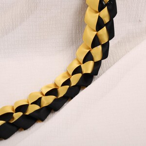 May include: A braided lei featuring alternating sections of gold and black satin ribbon. The lei is displayed against a white textured background, showcasing the woven pattern and color contrast. This handcrafted item is suitable for celebrations.