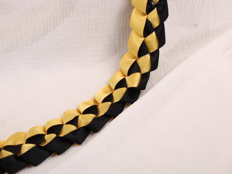 May include: A braided lei featuring alternating sections of gold and black satin ribbon. The lei is displayed against a white textured background, showcasing the woven pattern and color contrast. This handcrafted item is suitable for celebrations.