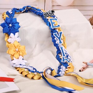 May include: A graduation lei in blue, yellow, and white, with the text "Class of 2026" and the name "EYIK". The lei features ribbon flowers and braided ribbon. A teddy bear is in the background.