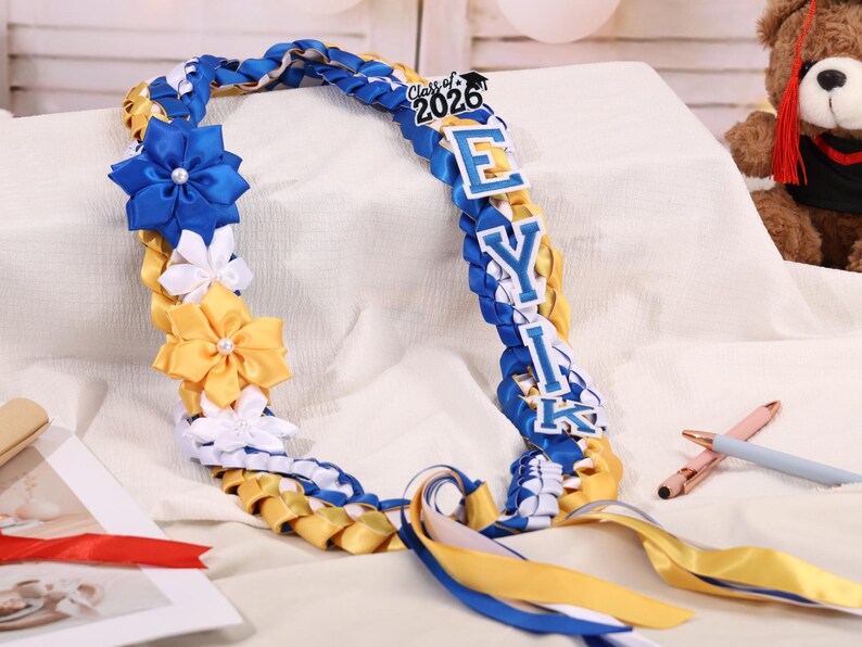 May include: A graduation lei in blue, yellow, and white, with the text "Class of 2026" and the name "EYIK". The lei features ribbon flowers and braided ribbon. A teddy bear is in the background.