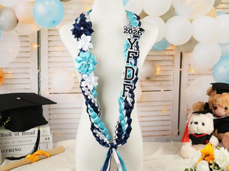 May include: A graduation lei in blue, white, and navy ribbons, adorned with fabric flowers and a "Class of 2026" charm. The lei features the letters "Y F D F" in white with a black outline. The lei is displayed on a mannequin.