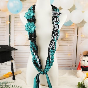May include: A graduation lei in teal, black, and white, with the name "DORA" spelled out in large letters. The lei is adorned with teal and black flower accents and a "Class of 2026" emblem. Ribbons hang from the bottom.