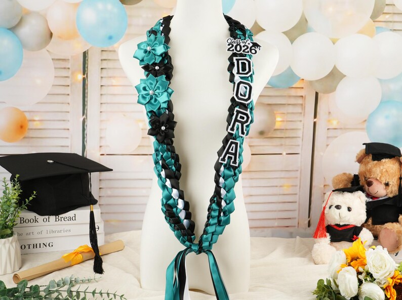 May include: A graduation lei in teal, black, and white, with the name "DORA" spelled out in large letters. The lei is adorned with teal and black flower accents and a "Class of 2026" emblem. Ribbons hang from the bottom.