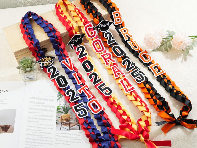 May include: Braided graduation leis in red, blue, yellow, and black. Each lei features the year "2025" and a name, with a graduation cap charm. A festive and personalized graduation gift.
