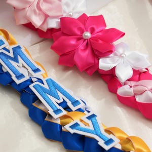 May include: Close-up of colorful leis, a traditional Hawaiian garland. The leis feature braided ribbons in blue, yellow, and pink, with white and pink satin flower accents. Some leis have the letters "IMMY" in blue and white.