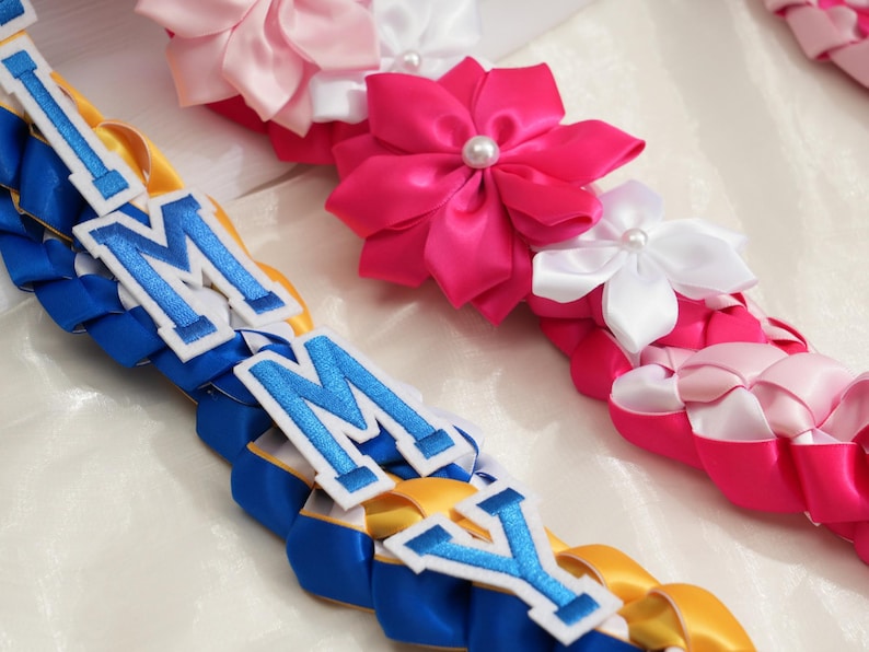 May include: Close-up of colorful leis, a traditional Hawaiian garland. The leis feature braided ribbons in blue, yellow, and pink, with white and pink satin flower accents. Some leis have the letters "IMMY" in blue and white.