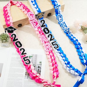 May include: Three graduation leis in pink, white, and blue. The pink lei has the text "Class of 2025". The white and pink lei has the name "GEAR" and the year "2025". The blue lei has the name "TONY".
