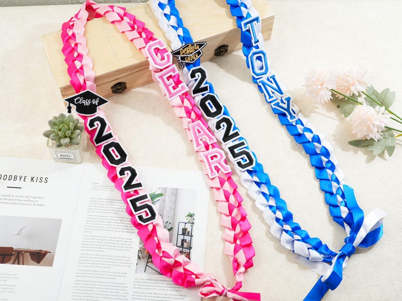 May include: Three graduation leis in pink, white, and blue. The pink lei has the text "Class of 2025". The white and pink lei has the name "GEAR" and the year "2025". The blue lei has the name "TONY".