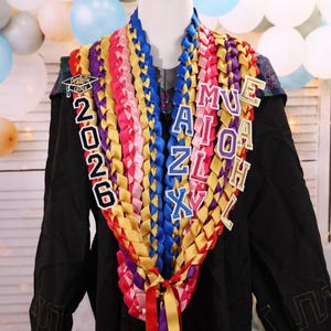 May include: A graduation lei with colorful ribbons in shades of red, pink, yellow, blue, and purple. The lei has the year "2026" and the name "MUEAIZLXH" in individual letter charms. The lei is displayed on a black graduation gown.