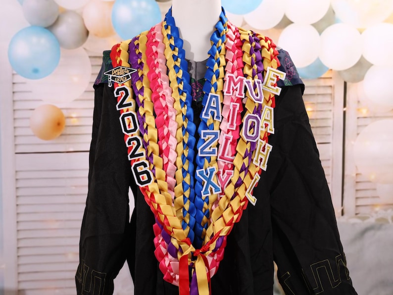 May include: A graduation lei with colorful ribbons in shades of red, pink, yellow, blue, and purple. The lei has the year "2026" and the name "MUEAIZLXH" in individual letter charms. The lei is displayed on a black graduation gown.