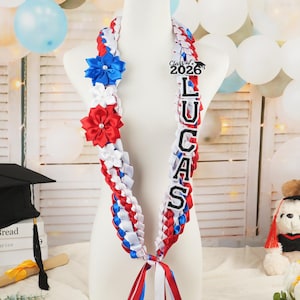 May include: A graduation lei in red, white, and blue ribbon, adorned with blue and red flower accents. The lei features the name "LUCAS" and "Class of 2026" on a black background. A festive graduation decoration.